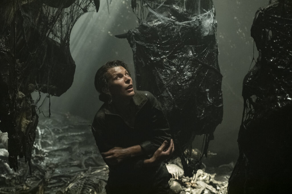 A young, white-passing woman looks around in fear, hugging herself surrounding by slimy, cocoon-like vessels hanging from above in a dark cavern.