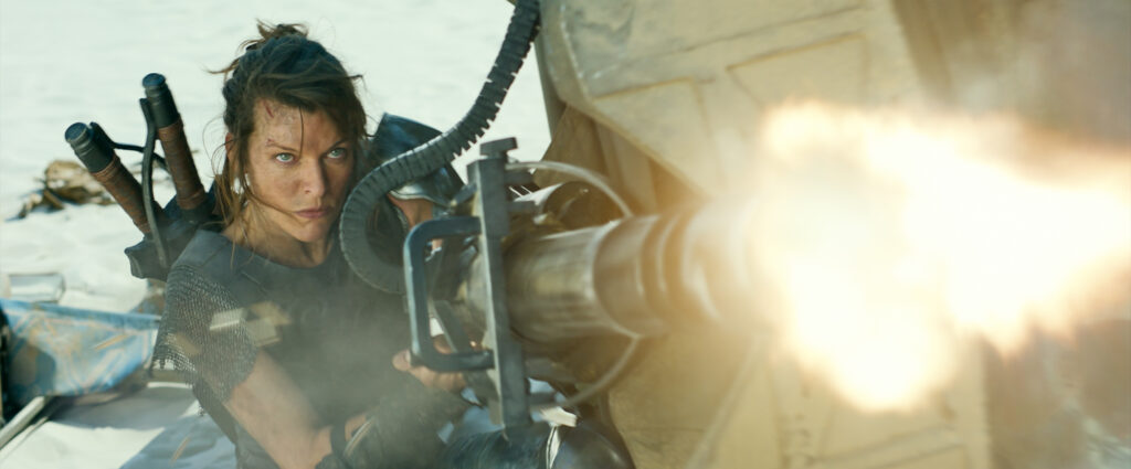 A young, white-passing woman shoots a machine gun with a fierce expression towards a direction off-frame.