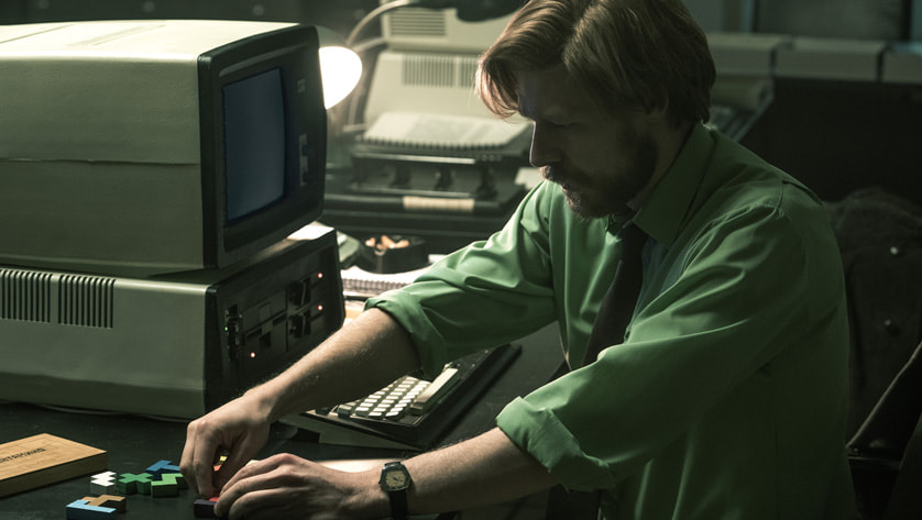 A man with a short-trimmed beard, and dark blonde, side-swept hair sits in front of a desk with an an old computer system. He wears a green collared dress shirt and black tie. He fiddles with differently colored blocks in front of him.